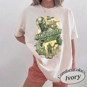 May include: Ivory-colored t-shirt featuring a graphic from The Mandalorian series. The design includes the text "The Mandalorian and Grogu" and "This is the Way." The shirt is a Comfort Colors brand.