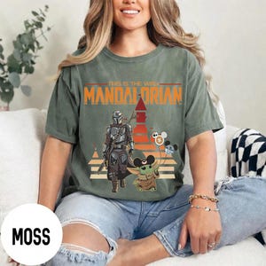 May include: Moss green t-shirt featuring a graphic of The Mandalorian and Grogu with the text "This is the Way" and "Mandalorian". The design includes a retro sunset and cartoon characters. The shirt is a comfortable, casual style.