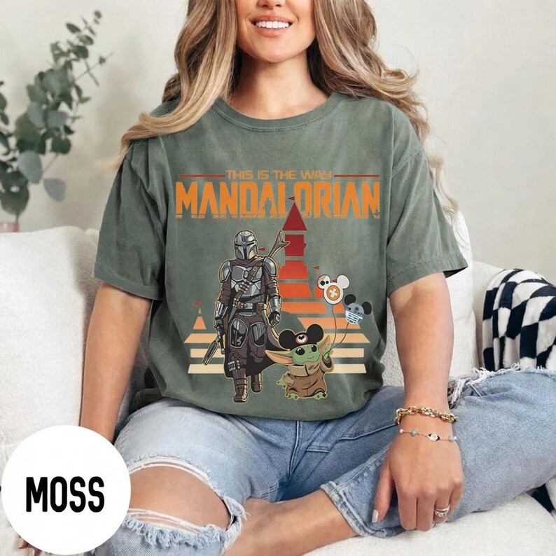 May include: Moss green t-shirt featuring a graphic of The Mandalorian and Grogu with the text "This is the Way" and "Mandalorian". The design includes a retro sunset and cartoon characters. The shirt is a comfortable, casual style.