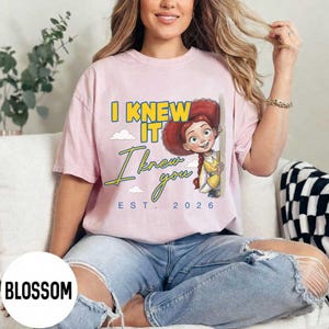May include: A light pink short-sleeve t-shirt featuring a graphic of the character Jessie from Toy Story. The text reads I KNEW IT in yellow, I knew you in blue script, and EST. 2026 in blue. The design includes small white clouds.