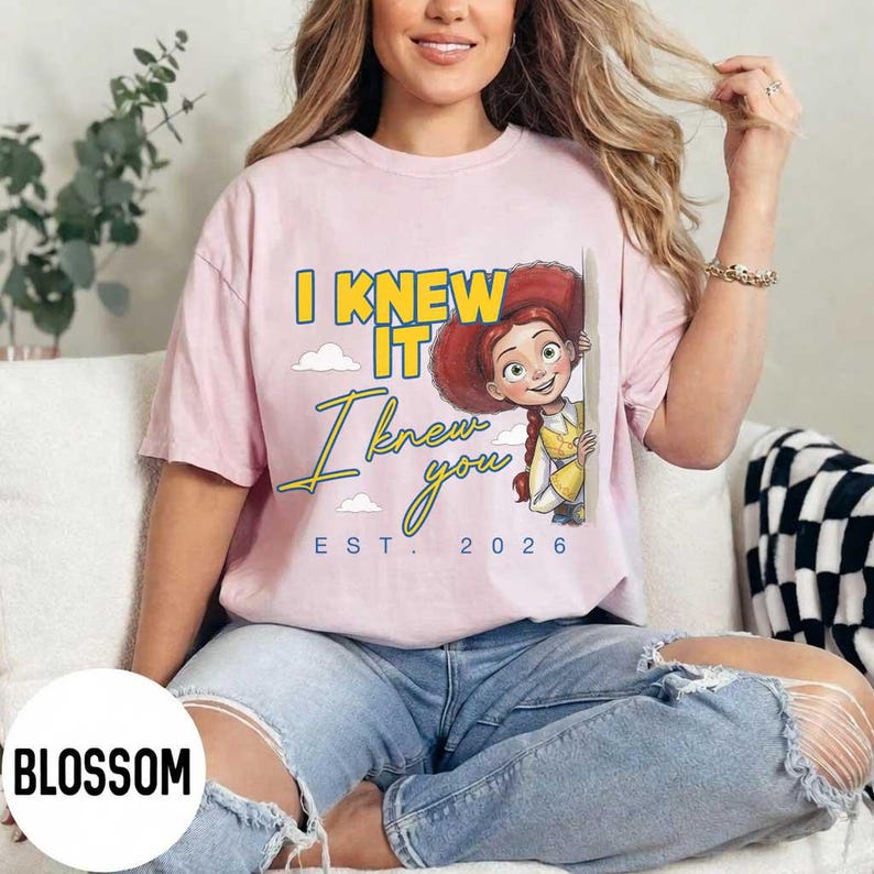 May include: A light pink short-sleeve t-shirt featuring a graphic of the character Jessie from Toy Story. The text reads I KNEW IT in yellow, I knew you in blue script, and EST. 2026 in blue. The design includes small white clouds.