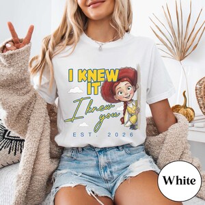 May include: A white short-sleeved t-shirt featuring a graphic of a red-haired cowgirl character peeking from behind a wall. The text reads I KNEW IT, I knew you, and EST. 2026 in yellow and blue lettering, accompanied by small white cloud illustrations.