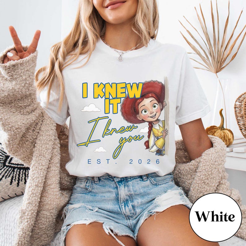 May include: A white short-sleeved t-shirt featuring a graphic of a red-haired cowgirl character peeking from behind a wall. The text reads I KNEW IT, I knew you, and EST. 2026 in yellow and blue lettering, accompanied by small white cloud illustrations.
