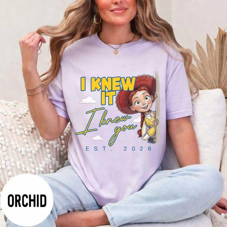 May include: A light purple t-shirt featuring a graphic of a cartoon cowgirl with red hair and a yellow shirt. The text reads I KNEW IT, I knew you, and EST. 2026 in yellow and blue lettering. The shirt has short sleeves and a crew neckline.