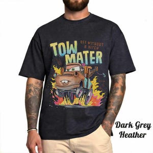 May include: A dark grey heather t-shirt featuring a graphic of the character Tow Mater from the movie Cars. The design includes the text "TOW MATER" and "OFF WITHOUT A HITCH" above the character, who is surrounded by stylized orange and yellow flames.