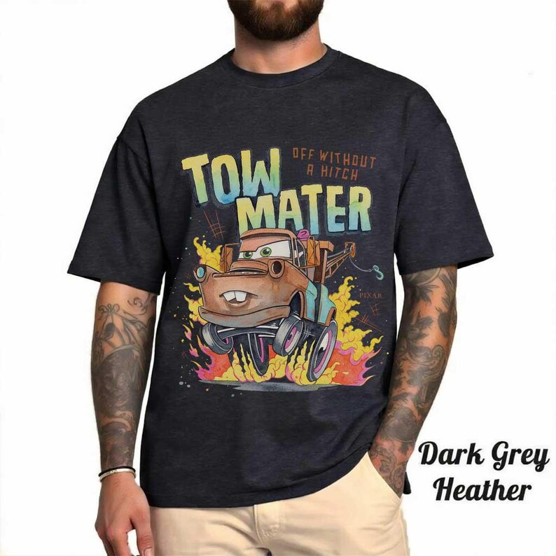 May include: A dark grey heather t-shirt featuring a graphic of the character Tow Mater from the movie Cars. The design includes the text "TOW MATER" and "OFF WITHOUT A HITCH" above the character, who is surrounded by stylized orange and yellow flames.