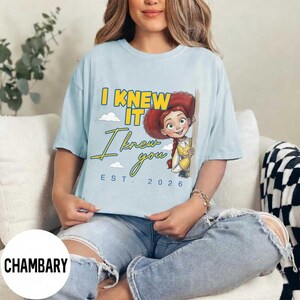 May include: A light blue t-shirt featuring a graphic of the character Jessie from Toy Story. The text reads I KNEW IT I knew you EST. 2026 in yellow and blue lettering. The design includes small white clouds and the character wearing a hat.