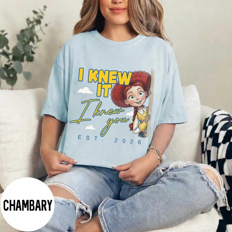 May include: A light blue t-shirt featuring a graphic of the character Jessie from Toy Story. The text reads I KNEW IT I knew you EST. 2026 in yellow and blue lettering. The design includes small white clouds and the character wearing a hat.