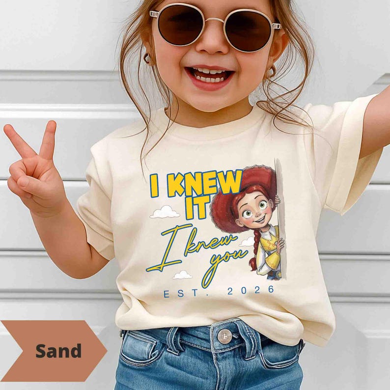 May include: A sand-colored t-shirt featuring a graphic of the character Jessie from Toy Story. The text reads I KNEW IT, I knew you, and EST. 2026 in yellow and blue lettering. The design includes small white clouds and a light blue border.