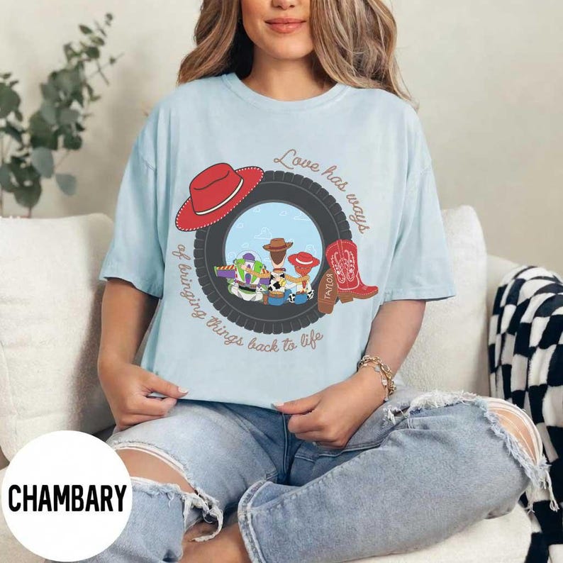 Pu&ograve; includere: Una t-shirt azzurro chiaro con la grafica di uno pneumatico nero che incornicia personaggi dei cartoni animati, tra cui un ranger spaziale e un cowboy. Il design include un cappello da cowboy rosso, uno stivale rosso e il testo Love has ways of bringing things back to life.