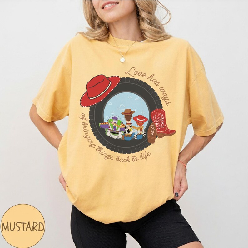 Pu&ograve; includere: Una t-shirt a maniche corte color giallo senape con la grafica di uno pneumatico nero che incornicia i personaggi di Toy Story. Il design include un cappello da cowboy rosso, uno stivale da cowboy rosso e il testo Love has ways of bringing things back to life.