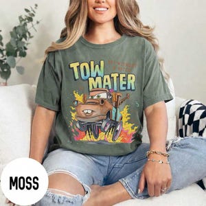 May include: A moss green short-sleeve t-shirt featuring a graphic of the character Tow Mater from the movie Cars. The text reads "TOW MATER" in large yellow and blue letters, with "OFF WITHOUT A HITCH" printed in smaller red text above it.
