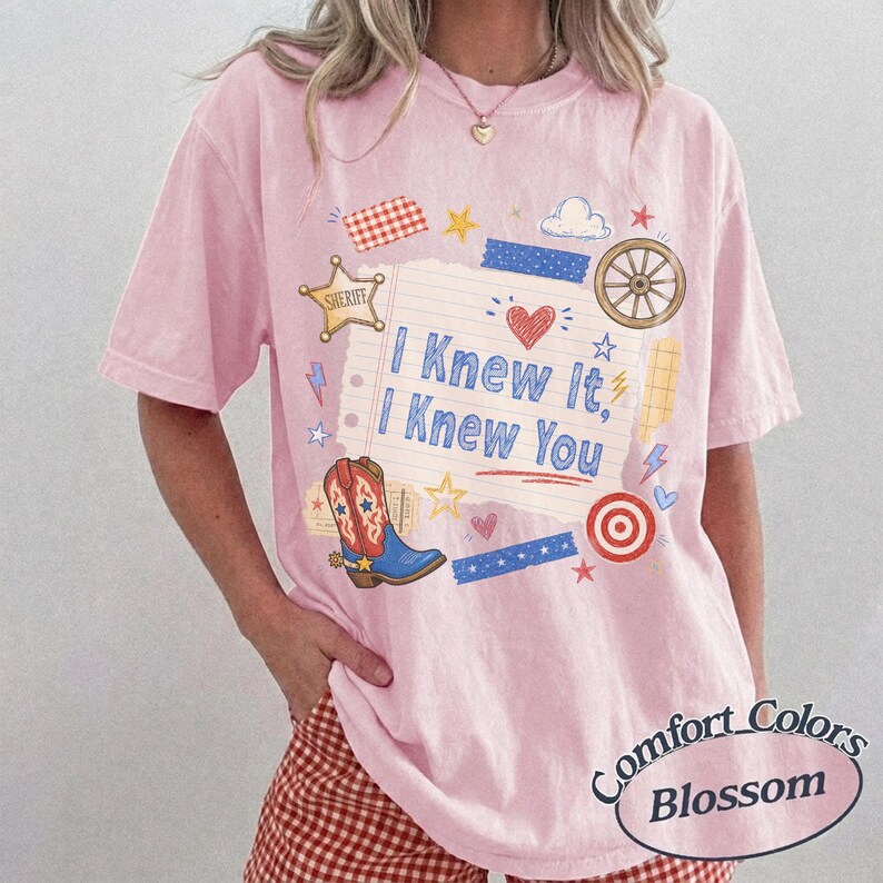 May include: A light pink short-sleeve t-shirt featuring a central graphic of a lined notebook paper with the text I Knew It, I Knew You. The design includes a sheriff star, a cowboy boot, a heart, a wagon wheel, and various colorful shapes.