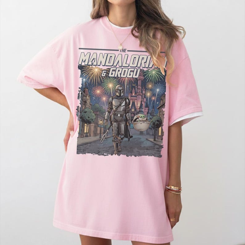 Op de afbeelding: Een lichtroze T-shirt met korte mouwen en een grafische print van The Mandalorian en Grogu. De illustratie toont de personages die over een straat lopen met een kasteel op de achtergrond onder een lucht vol met kleurrijke vuurwerken.