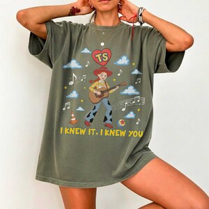 May include: An olive green t-shirt featuring a graphic of a cowgirl playing an acoustic guitar. The design includes a red heart with the letters TS, musical notes, clouds, a traffic cone, a ball, and the yellow text I KNEW IT, I KNEW YOU.