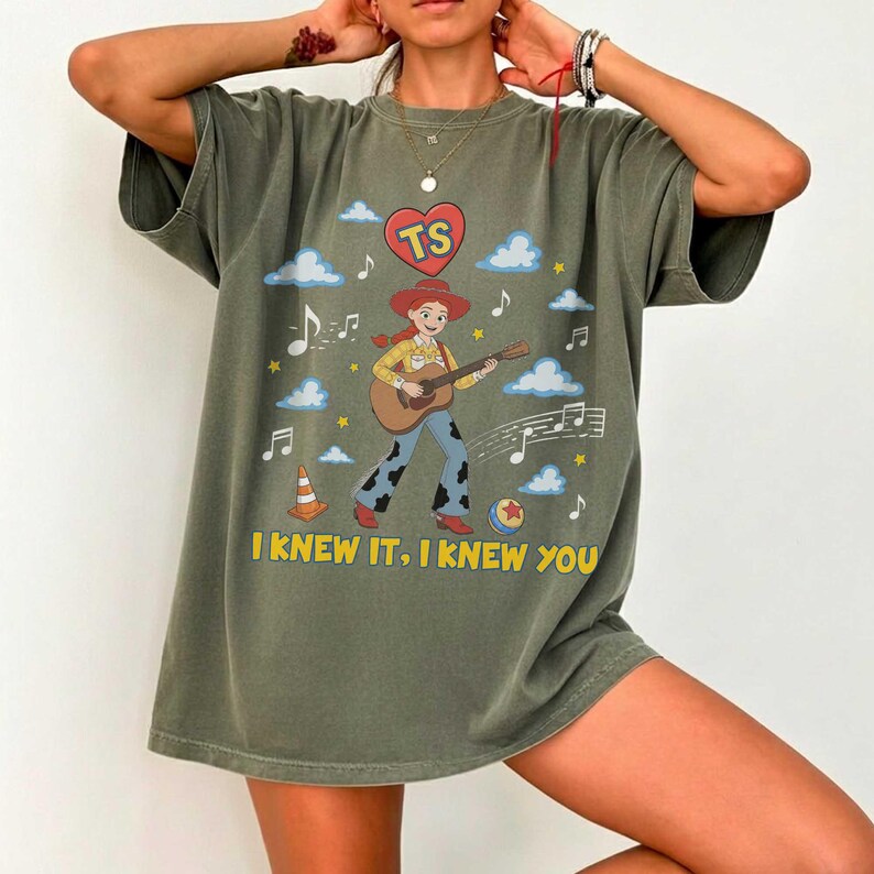 May include: An olive green t-shirt featuring a graphic of a cowgirl playing an acoustic guitar. The design includes a red heart with the letters TS, musical notes, clouds, a traffic cone, a ball, and the yellow text I KNEW IT, I KNEW YOU.