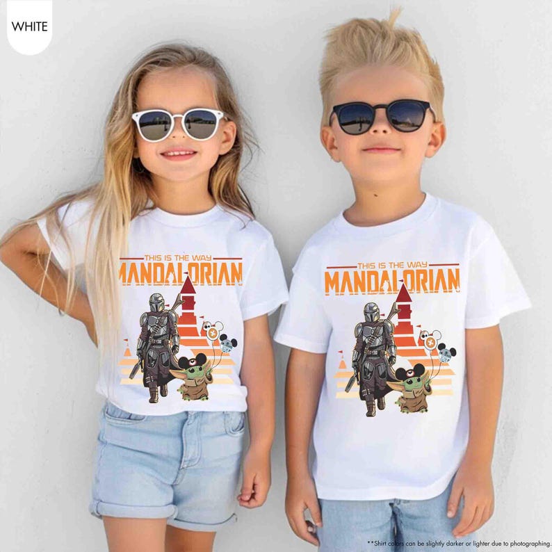 May include: Two children wearing white t-shirts with a "Mandalorian" graphic. The shirts feature the text "This is the Way" above the graphic of a Mandalorian character, Grogu, and other design elements. The children are wearing sunglasses.