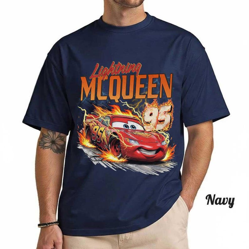May include: A navy blue t-shirt featuring a graphic of the red race car Lightning McQueen surrounded by orange and yellow flames. The text Lightning MCQUEEN is printed in orange above the car, with the number 95 displayed on the car and side.