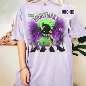Pu&ograve; includere: Una maglietta lilla chiaro con una grafica di "Nightmare Before Christmas". Il design include il testo "The Nightmare" sopra un'immagine verde e viola di Jack Skellington e altri personaggi. La maglietta ha maniche corte.