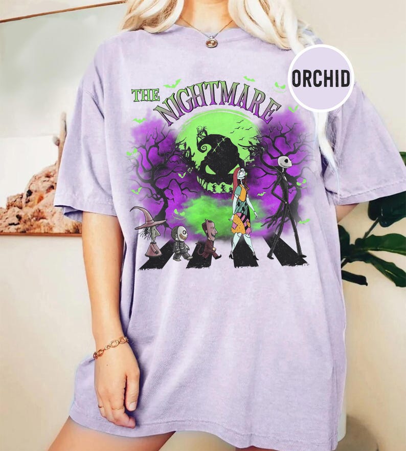 Pu&ograve; includere: Una maglietta lilla chiaro con una grafica di "Nightmare Before Christmas". Il design include il testo "The Nightmare" sopra un'immagine verde e viola di Jack Skellington e altri personaggi. La maglietta ha maniche corte.