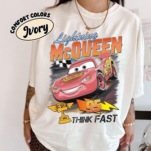 May include: Ivory-colored t-shirt featuring a graphic of Lightning McQueen from the movie Cars. The design includes the text "Lightning McQueen", "95", and "Think Fast". The shirt also has the words "Comfort Colors" and "Ivory" printed on it.