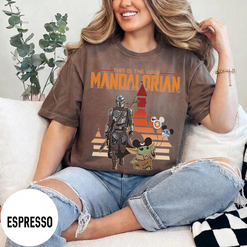 May include: Espresso-colored t-shirt featuring a graphic of The Mandalorian and Grogu, with the text "THIS IS THE WAY MANDALORIAN". The design includes a retro sunset and Mickey Mouse-themed balloons. The shirt is worn by a person.