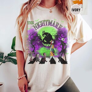 Pu&ograve; includere: T-shirt color avorio con grafica di "Nightmare Before Christmas". Il design include personaggi come Jack Skeletron e Sally, con una luna verde e alberi viola. Il testo "The Nightmare" &egrave; arcuato nella parte superiore.