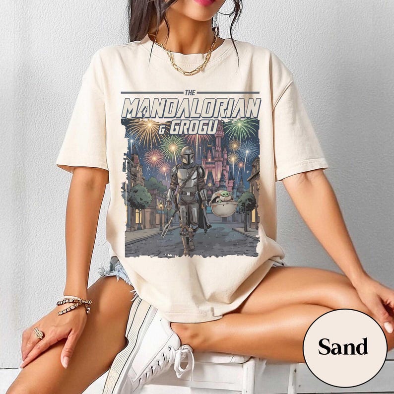 Op de afbeelding: Een zandkleurig t-shirt met een grafische print van The Mandalorian en Grogu die over een straat lopen. Erboven staat de tekst THE MANDALORIAN & GROGU. De achtergrond toont een kasteel verlicht door kleurrijk vuurwerk in de nachtelijke hemel.
