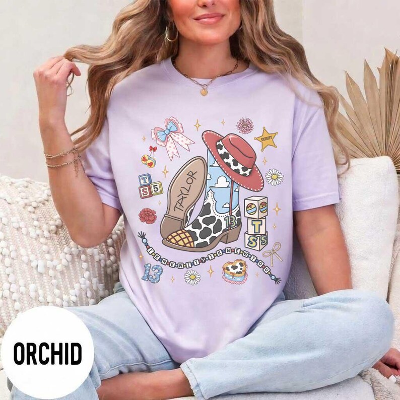 Op de afbeelding: Een lichtpaars t-shirt met een grafische print van een cowboylaars met koeienprint, een rode cowboyhoed, een blauwe strik, alfabetblokken met de letters T, S en 13, en de tekst Taylor. Het ontwerp bevat sterren en bloemen.