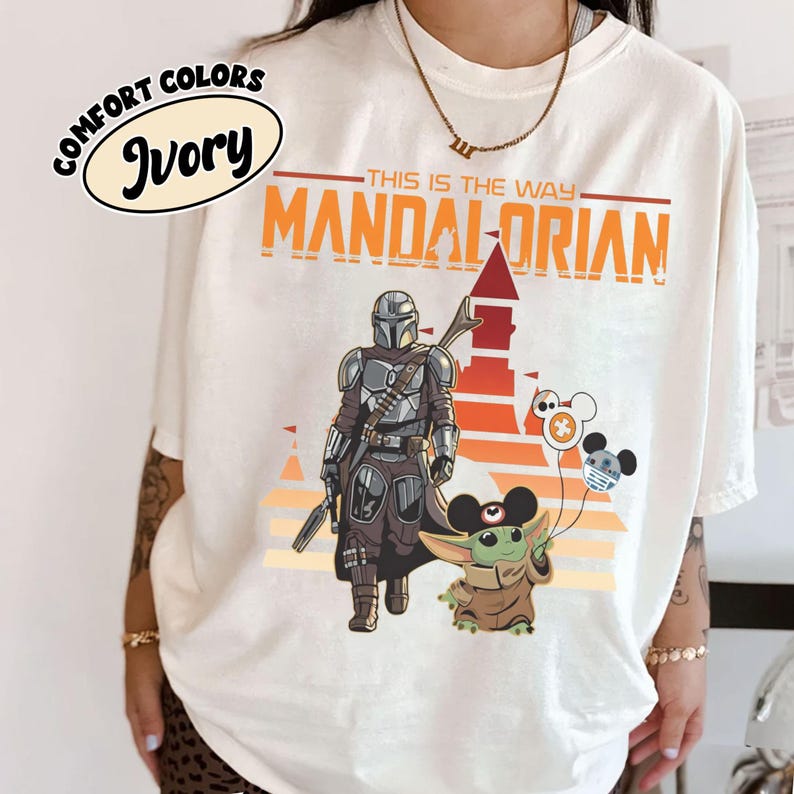 May include: Ivory Comfort Colors t-shirt featuring a graphic of The Mandalorian and Grogu. The design includes the text "This is the Way" and a stylized sunset background. Grogu holds Mickey Mouse balloons.