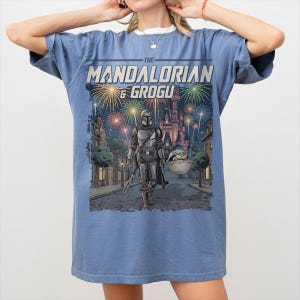 Op de afbeelding: Een blauw T-shirt met korte mouwen en een grafische print van The Mandalorian en Grogu die over een straat lopen, met vuurwerk en een kasteel op de achtergrond. De tekst "THE MANDALORIAN & GROGU" staat in een vetgedrukt lettertype boven de sc&egrave;ne.