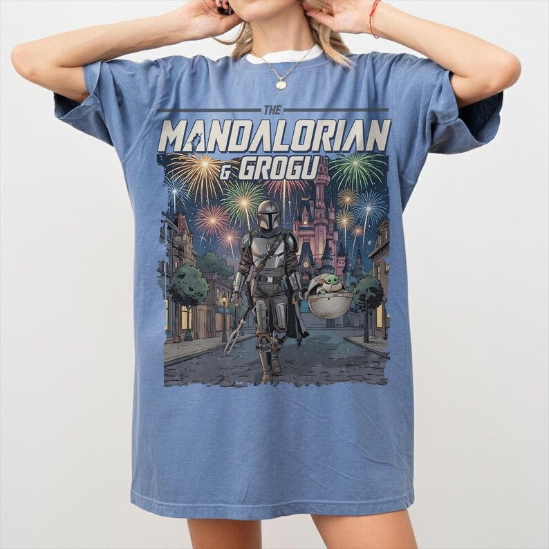 Op de afbeelding: Een blauw T-shirt met korte mouwen en een grafische print van The Mandalorian en Grogu die over een straat lopen, met vuurwerk en een kasteel op de achtergrond. De tekst "THE MANDALORIAN & GROGU" staat in een vetgedrukt lettertype boven de sc&egrave;ne.