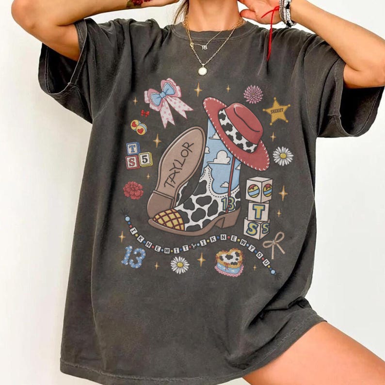 Op de afbeelding: Een antracietgrijs t-shirt met korte mouwen en een grafische print van een cowboylaars met koeienprint, een rode hoed, een strik, alfabetblokken met de letters T, S en 5, een hartvormige taart en de tekst Taylor, 13 en I KNEW IT I KNEW YOU.