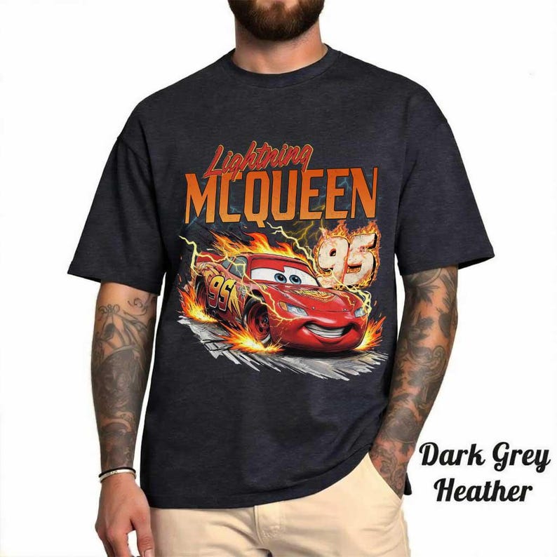 May include: A dark grey heather t-shirt featuring a graphic of the red race car Lightning McQueen surrounded by orange and yellow flames. The text Lightning MCQUEEN and the number 95 are printed above the car in bold orange lettering.