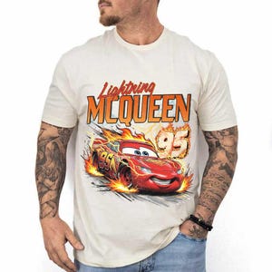 May include: A cream-colored short-sleeve t-shirt featuring a graphic of the red race car Lightning McQueen surrounded by orange flames. The text Lightning MCQUEEN is printed in orange and red above the car, with the number 95 in orange.
