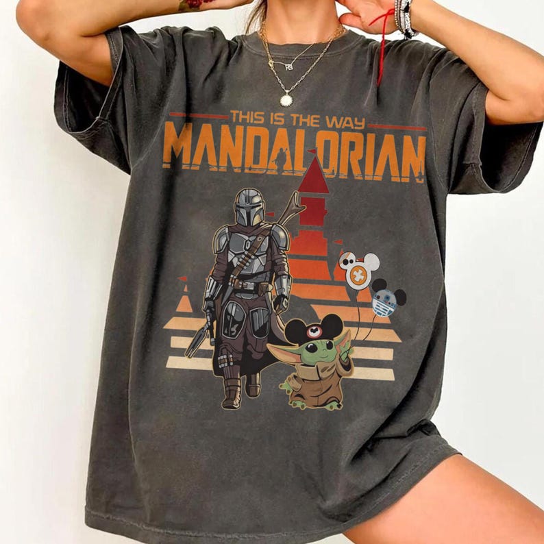 May include: Dark gray t-shirt featuring a graphic of The Mandalorian and Grogu from the Star Wars series. The design includes the text "THIS IS THE WAY" and "MANDALORIAN" in orange and white. The shirt also has Mickey Mouse-themed characters.