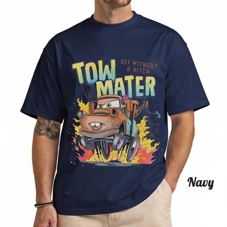 May include: A navy blue short-sleeve t-shirt featuring a graphic of the character Tow Mater from the movie Cars. The design includes the text "TOW MATER" and "OFF WITHOUT A HITCH" above the character, surrounded by stylized orange and yellow flames.