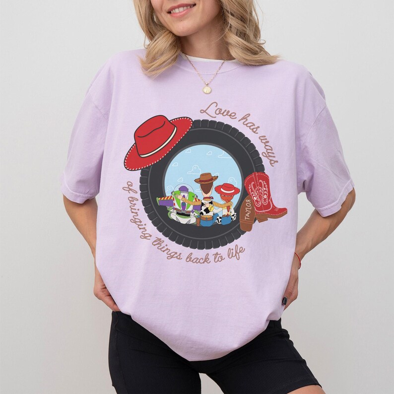 Pu&ograve; includere: Una t-shirt a maniche corte color viola chiaro con la grafica di uno pneumatico nero che incornicia i personaggi dei cartoni animati Buzz Lightyear, Woody e Jessie. Il design include un cappello da cowboy rosso, uno stivale rosso e il testo Love has ways of bringing things back to life.