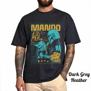 May include: A dark grey heather t-shirt featuring a graphic print of Mando and Grogu. The design includes the text MANDO AND GROGU, WHEREVER I GO, HE GOES, and THE LEGEND CONTINUES, along with illustrations of the characters in blue and gold tones.