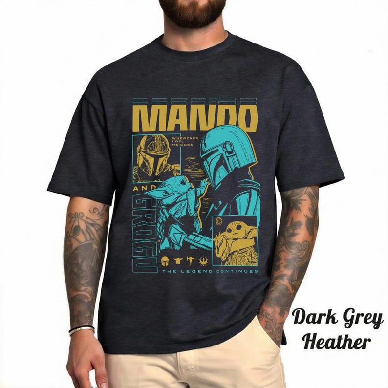 May include: A dark grey heather t-shirt featuring a graphic print of Mando and Grogu. The design includes the text MANDO AND GROGU, WHEREVER I GO, HE GOES, and THE LEGEND CONTINUES, along with illustrations of the characters in blue and gold tones.