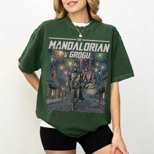 Op de afbeelding: Een donkergroen t-shirt met korte mouwen en een grafische print van The Mandalorian en Grogu die over een straat lopen met vuurwerk en een kasteel op de achtergrond. De tekst THE MANDALORIAN & GROGU staat in het wit boven de illustratie.