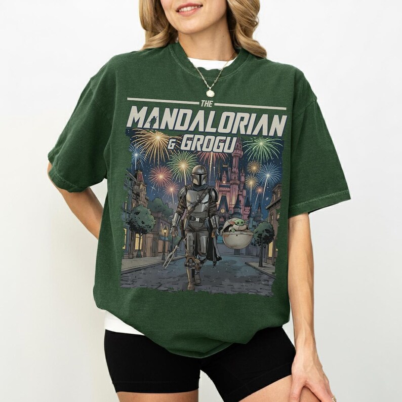 Op de afbeelding: Een donkergroen t-shirt met korte mouwen en een grafische print van The Mandalorian en Grogu die over een straat lopen met vuurwerk en een kasteel op de achtergrond. De tekst THE MANDALORIAN & GROGU staat in het wit boven de illustratie.