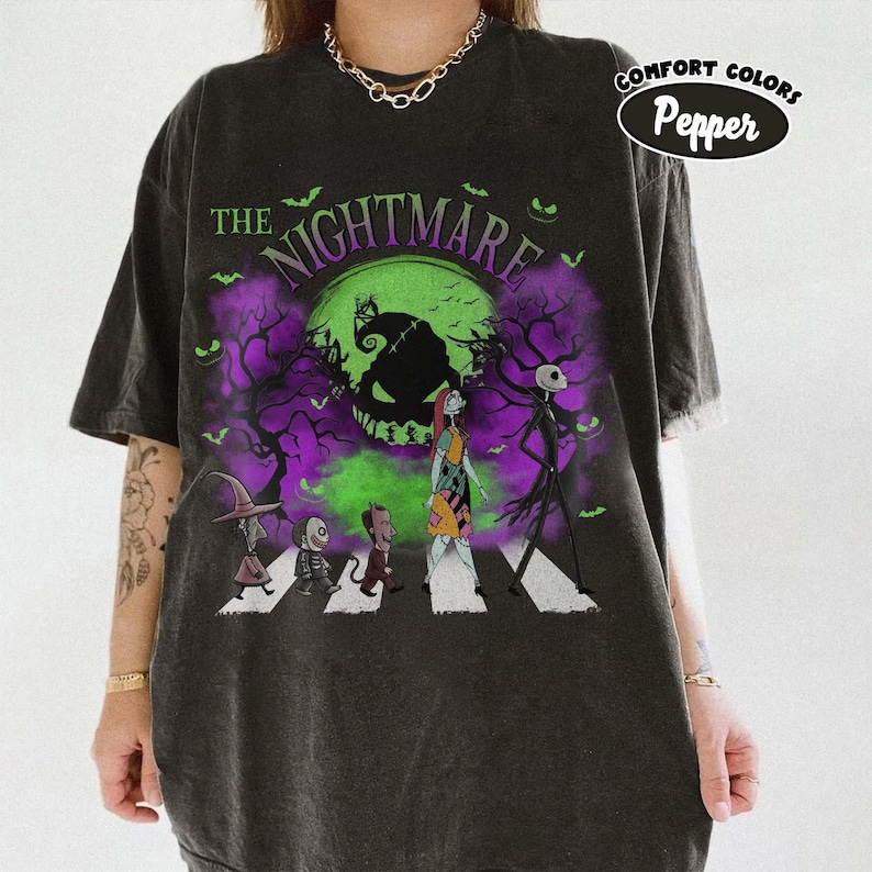 Pu&ograve; includere: T-shirt grigio scuro con grafica di "Nightmare Before Christmas". Il design mostra personaggi che attraversano una strada con una luna verde e accenti viola. Il testo "THE NIGHTMARE" &egrave; arcuato nella parte superiore.