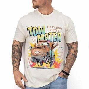 May include: A cream-colored short-sleeve t-shirt featuring a graphic of the character Tow Mater from the movie Cars. The design includes the text "TOW MATER" and "OFF WITHOUT A HITCH" in a colorful font, with flames at the bottom and the Pixar logo.