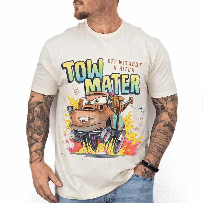 May include: A cream-colored short-sleeve t-shirt featuring a graphic of the character Tow Mater from the movie Cars. The design includes the text "TOW MATER" and "OFF WITHOUT A HITCH" in a colorful font, with flames at the bottom and the Pixar logo.
