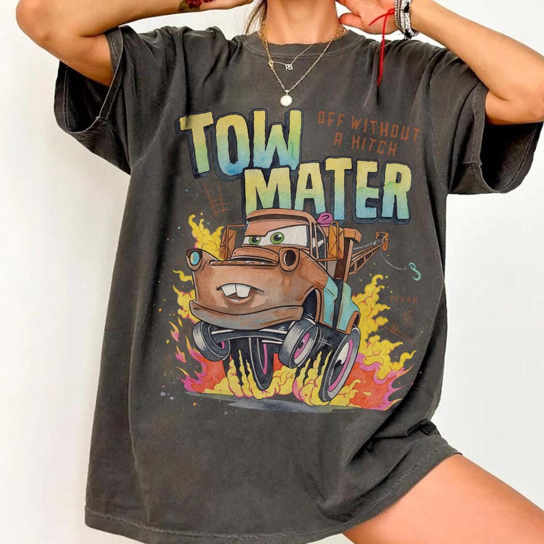 May include: A charcoal gray short-sleeve t-shirt featuring a graphic print of the character Tow Mater from the movie Cars. The text reads TOW MATER and OFF WITHOUT A HITCH. The design includes a brown tow truck surrounded by yellow and orange flames.