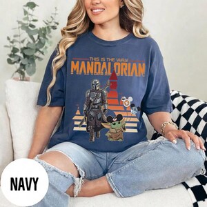 May include: Navy blue t-shirt featuring a graphic of The Mandalorian and Grogu with the text "THIS IS THE WAY MANDALORIAN". The design includes a sunset-colored background and Mickey Mouse-themed balloons.