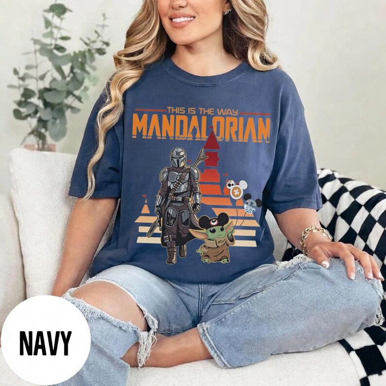 May include: Navy blue t-shirt featuring a graphic of The Mandalorian and Grogu with the text "THIS IS THE WAY MANDALORIAN". The design includes a sunset-colored background and Mickey Mouse-themed balloons.