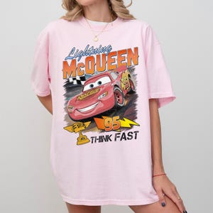May include: A light pink t-shirt featuring a graphic of Lightning McQueen from the movie Cars. The design includes the text "Lightning McQueen" and "95 THINK FAST". The shirt has short sleeves.