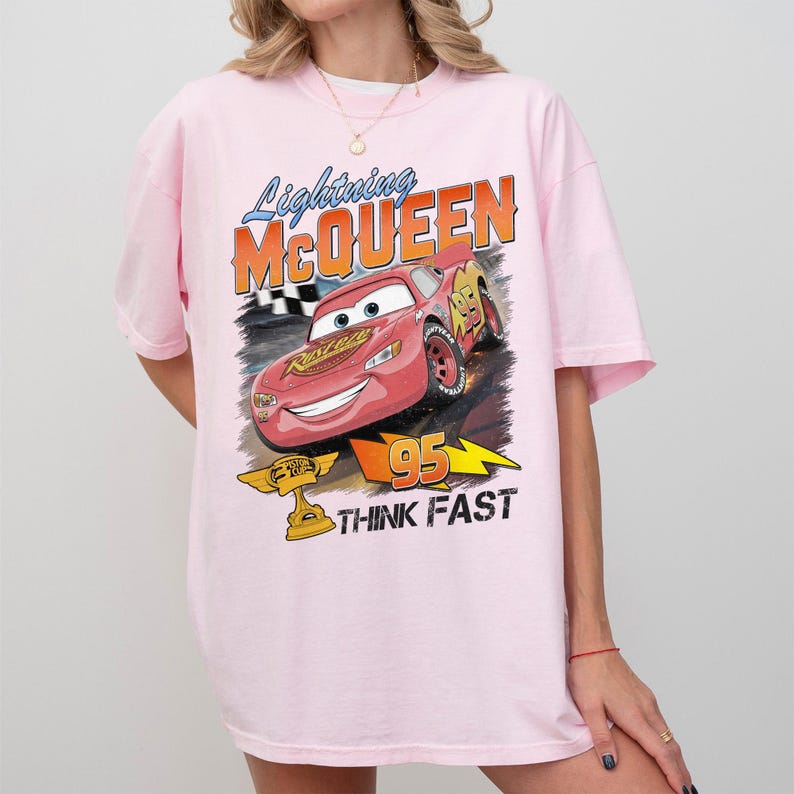 May include: A light pink t-shirt featuring a graphic of Lightning McQueen from the movie Cars. The design includes the text "Lightning McQueen" and "95 THINK FAST". The shirt has short sleeves.